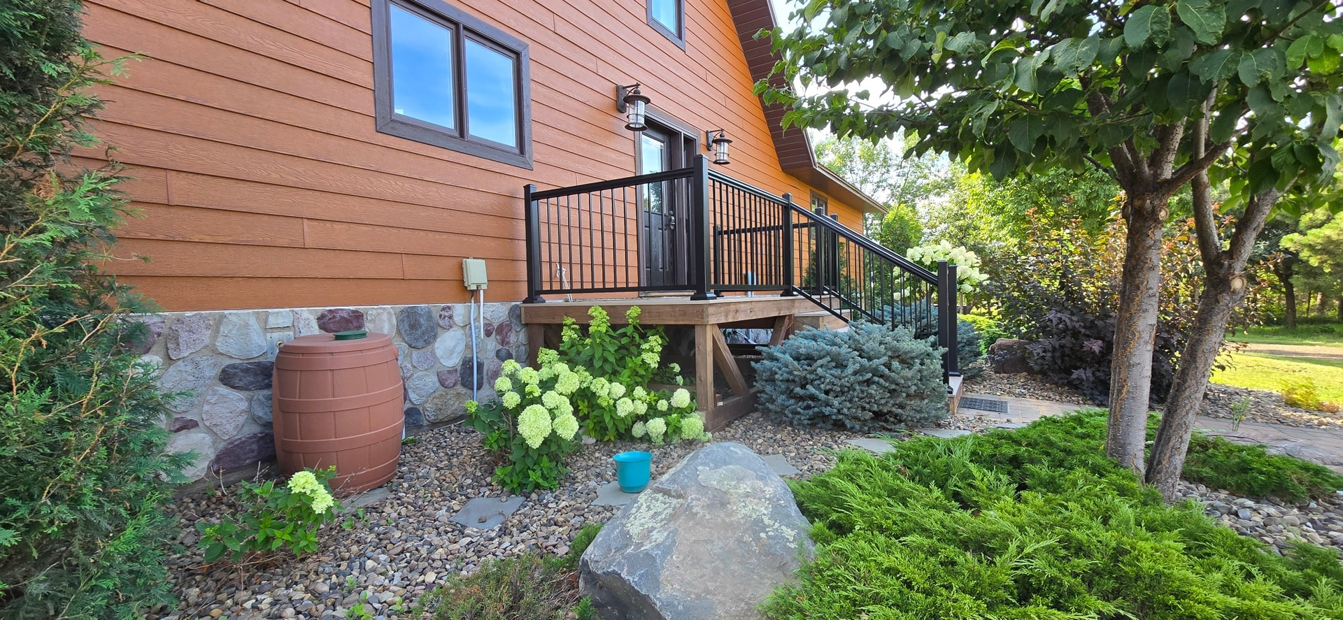 A brown wooden house with a stone foundation, a wooden deck, a rain barrel, and landscaping, set in a yard with trees.