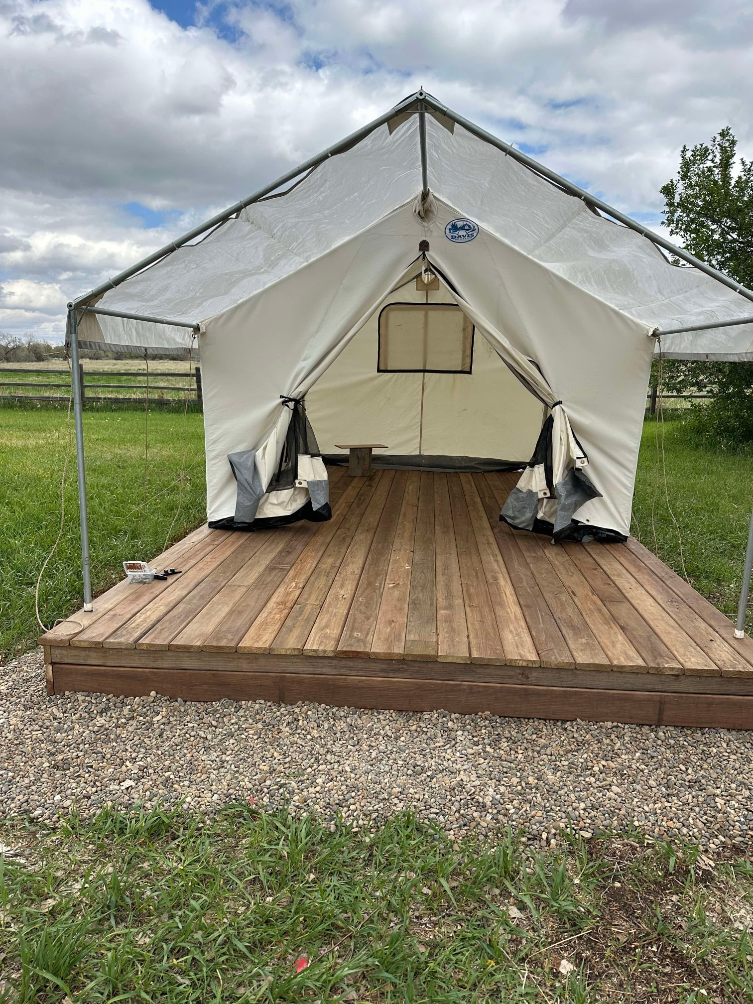 A white canvas wall tent on a wooden deck platform, set in a grassy field under a cloudy sky.