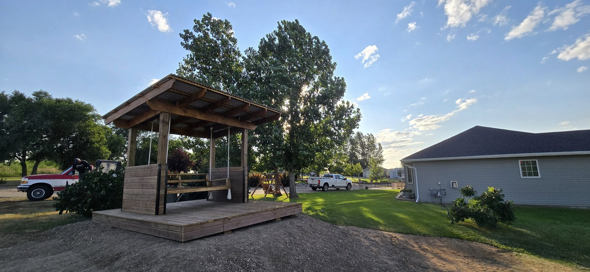 A wooden gazebo with a stone base and swing stands on a gravel yard next to a house and green trees under a sunny sky.