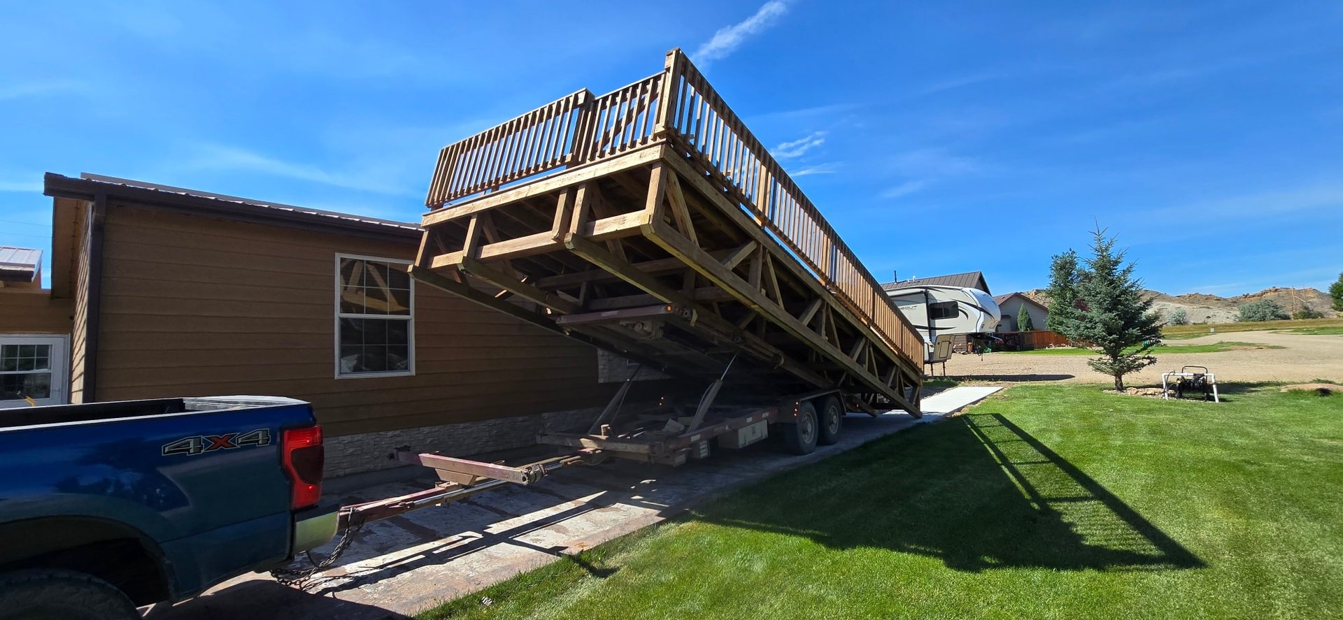 A wooden deck structure is being transported on a trailer towed by a blue pickup truck in a sunny residential yard.