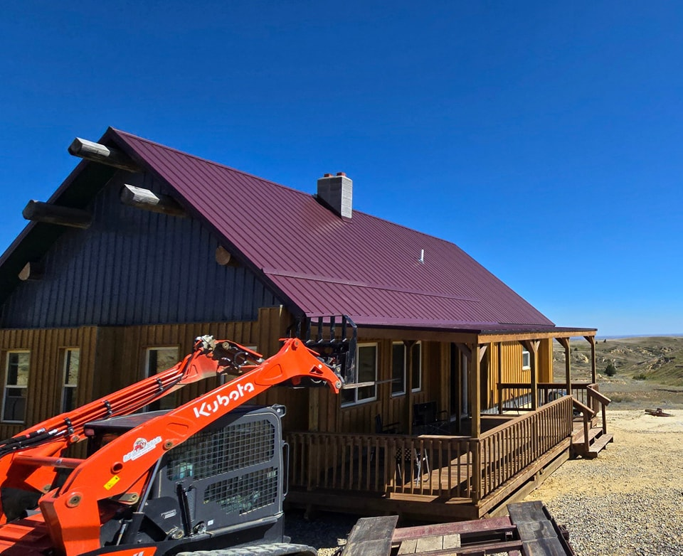An orange skid steer loader parked next to a house with a burgundy metal roof and a wooden deck under a clear blue sky.