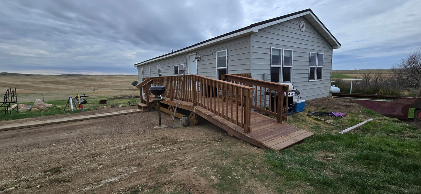 A light-gray mobile home sits on a grassy prairie with a wooden wheelchair ramp leading to the entrance.