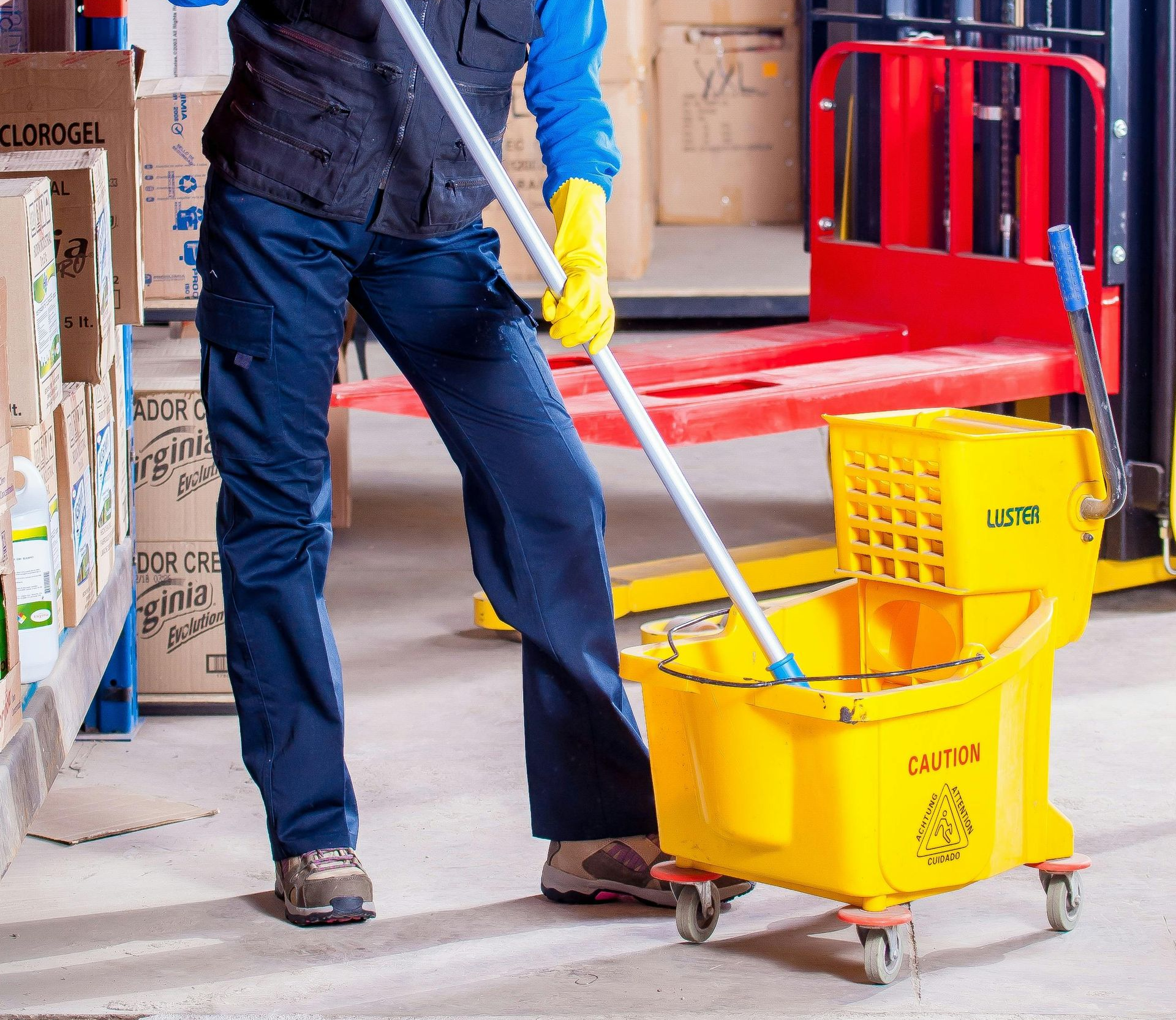 Person mopping a warehouse floor with a yellow mop bucket and wearing gloves.