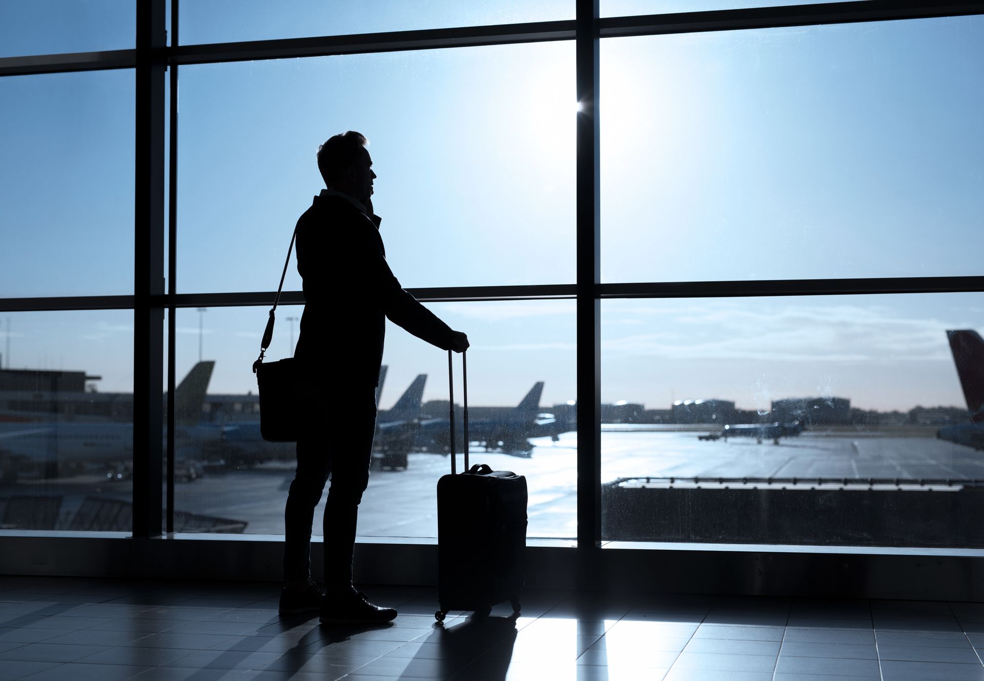 Silhouette of a traveler with suitcase at an airport window, looking toward airplanes on the tarmac.