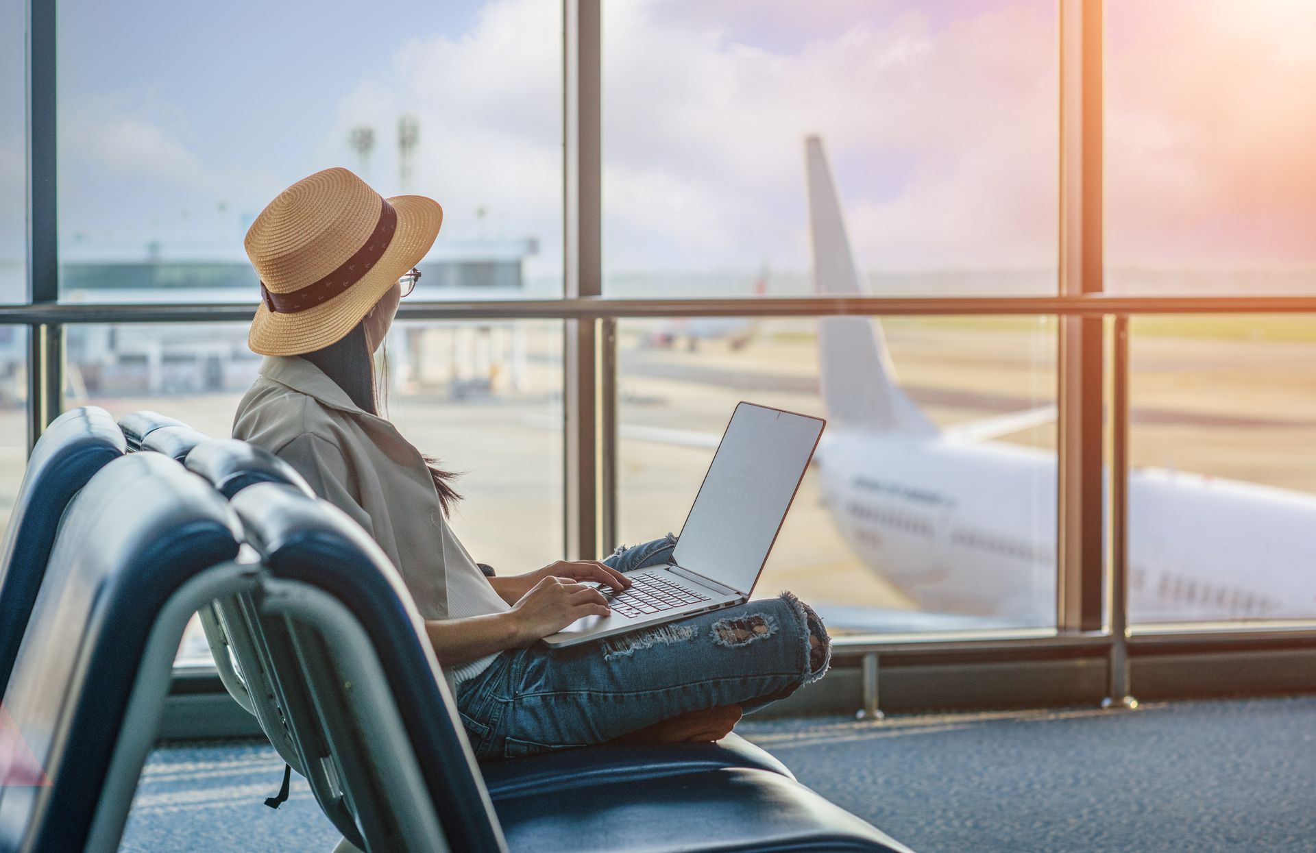 Traveler using a laptop by airport window, watching a plane outside at sunset
