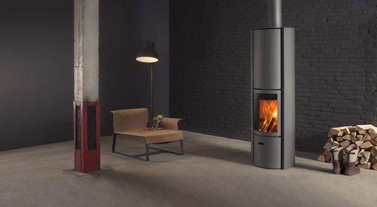 Moderna stufa con fuoco acceso accanto a una catasta di legna, una sedia e una lampada in una stanza grigia.