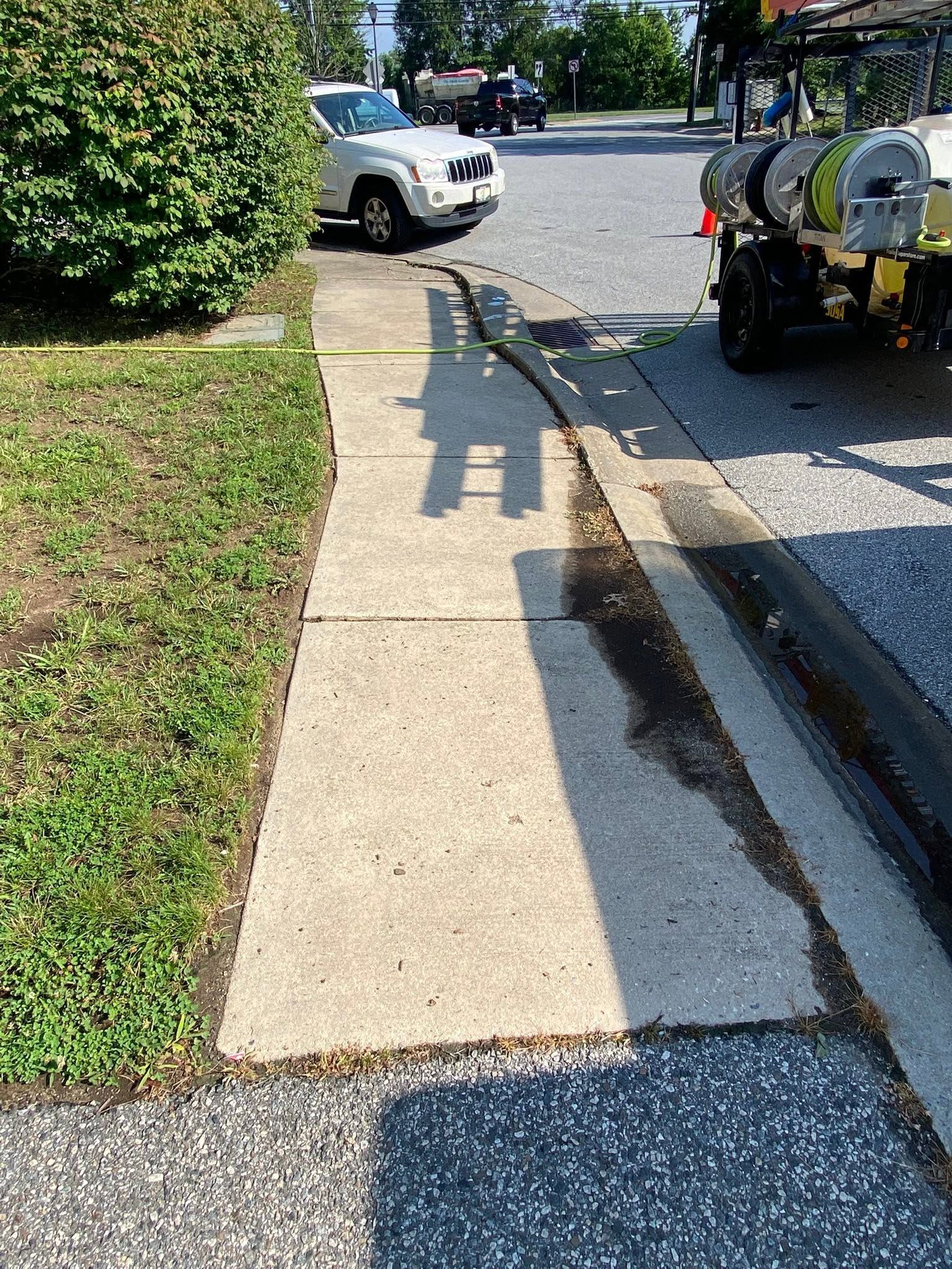Sidewalk next to a road with a parked utility truck, white car, and a hose casting a long shadow.