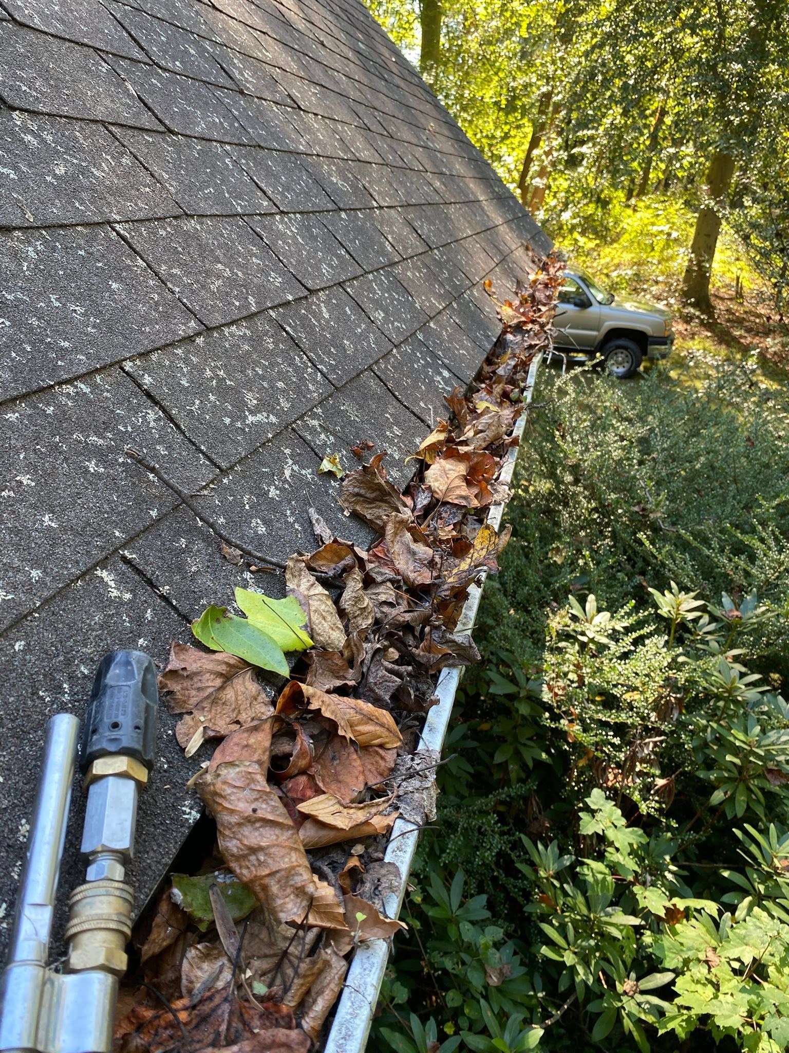 Gutter full of wet leaves and debris along a shingled roof, with tools on the left.