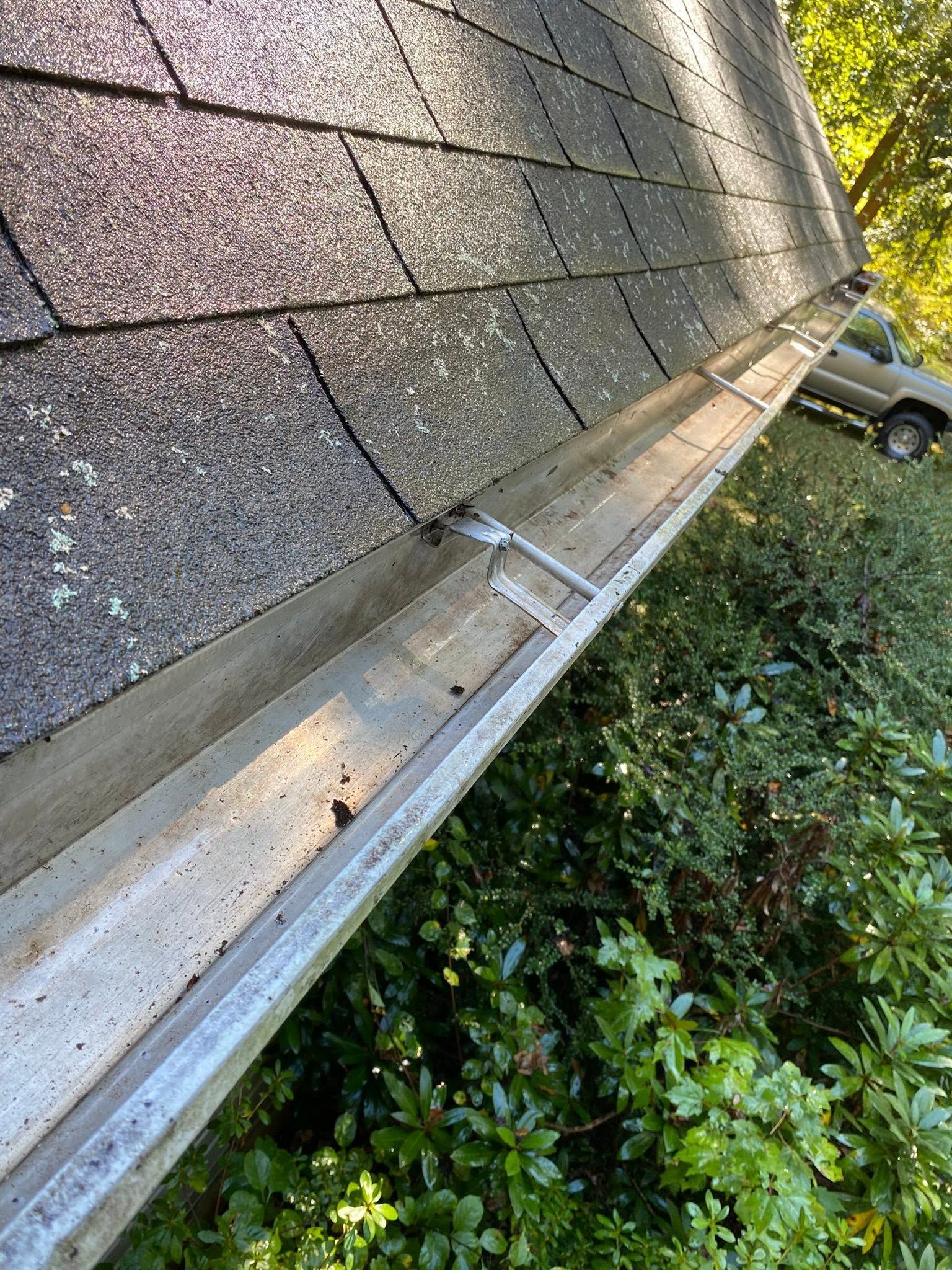 Roof gutter clogged with debris beside shingled roof and bushes near a parked car