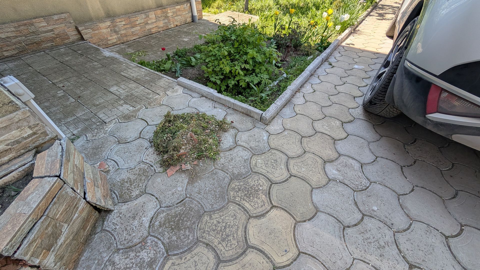 Broken hexagonal sidewalk beside a car, with weeds growing in cracks and a small step to a paved area
