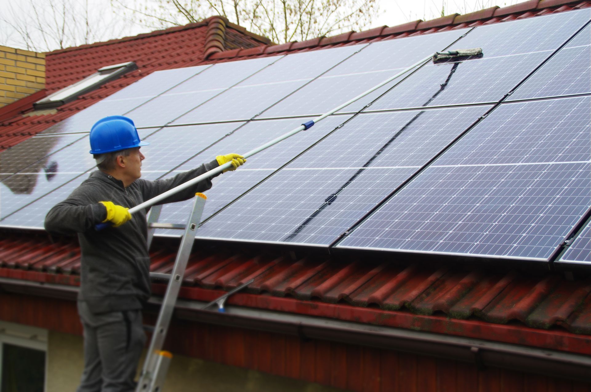 Worker cleaning rooftop solar panels with a hose on a red-tiled house roof