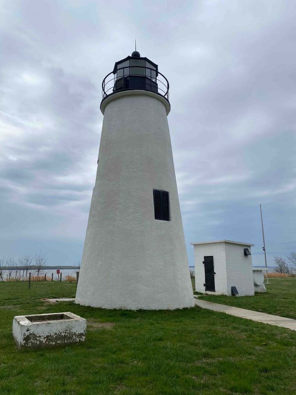 White lighthouse on grassy coast under cloudy sky, with a small white utility building beside it