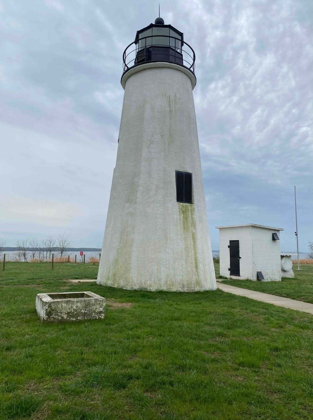 White lighthouse with black lantern beside a small white outbuilding on a grassy coast under cloudy sky