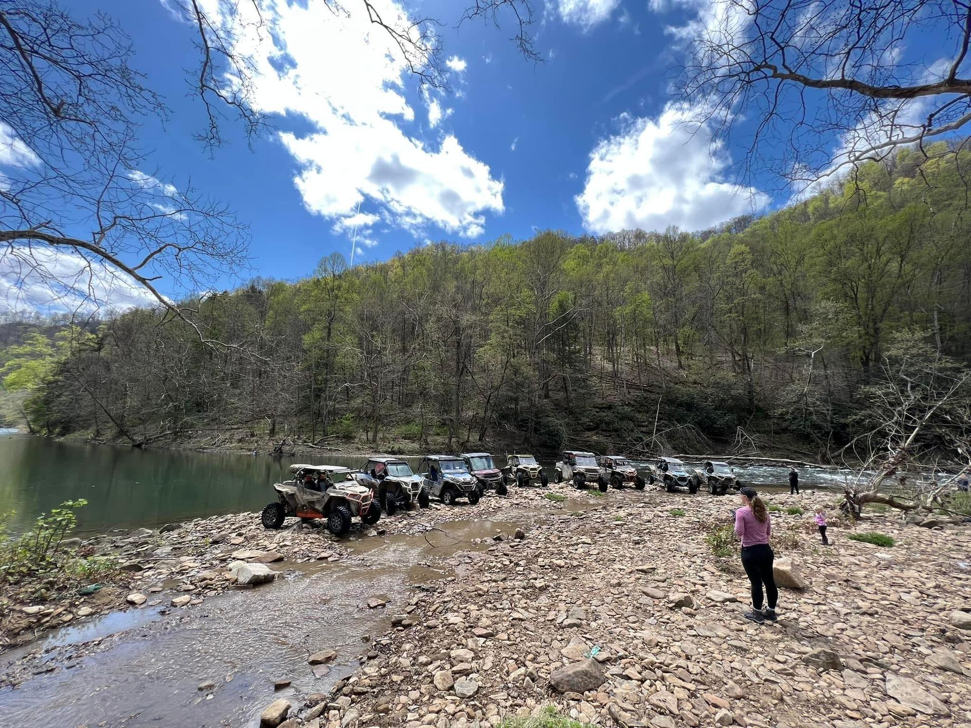 Off-road vehicles parked on a rocky riverbank under a bright blue sky, with one person standing nearby