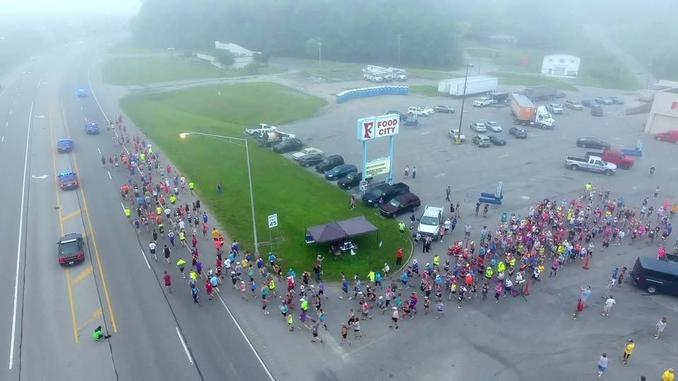 Large crowd in colorful shirts gathered in a foggy parking lot beside a road and grassy median.