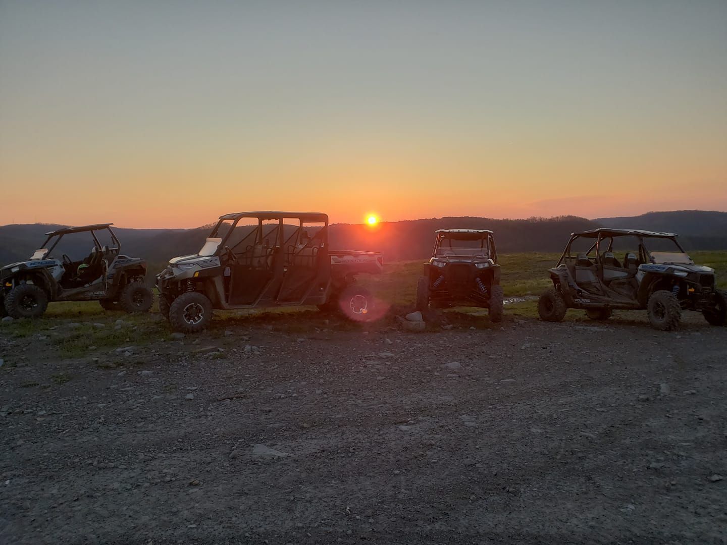 Four off-road vehicles parked at sunset on a gravel lot, silhouetted against an orange sky.
