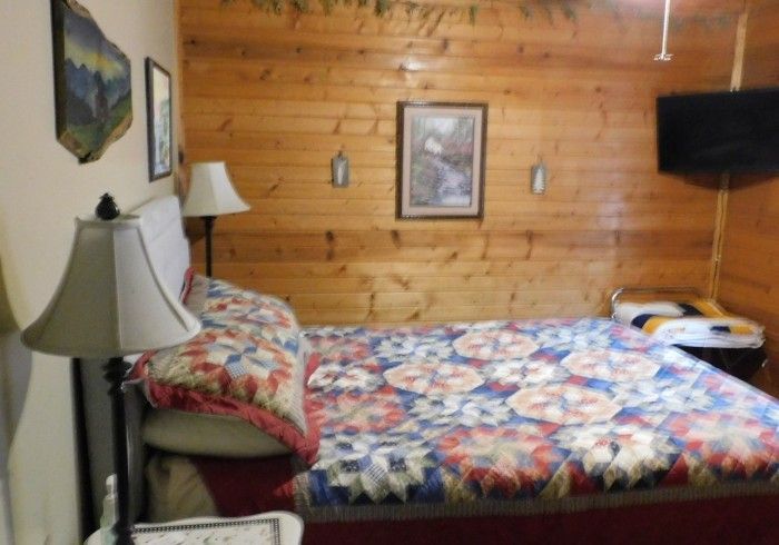 Cozy wood-paneled bedroom with a floral bedspread, lamp, framed art, and wall-mounted TV.