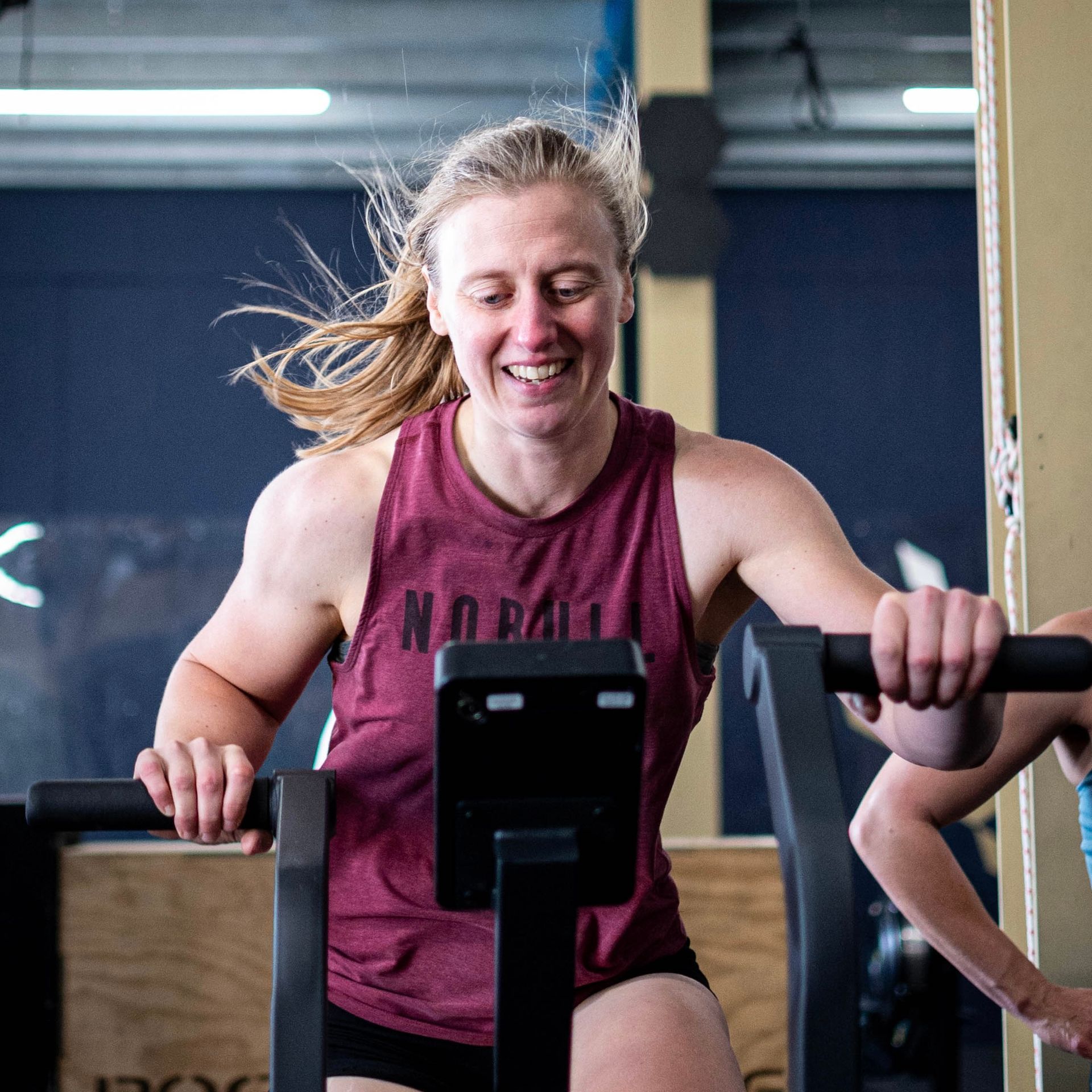 Vrouw in een bordeauxrode tanktop op een hometrainer, lachend en sportend in een sportschool.