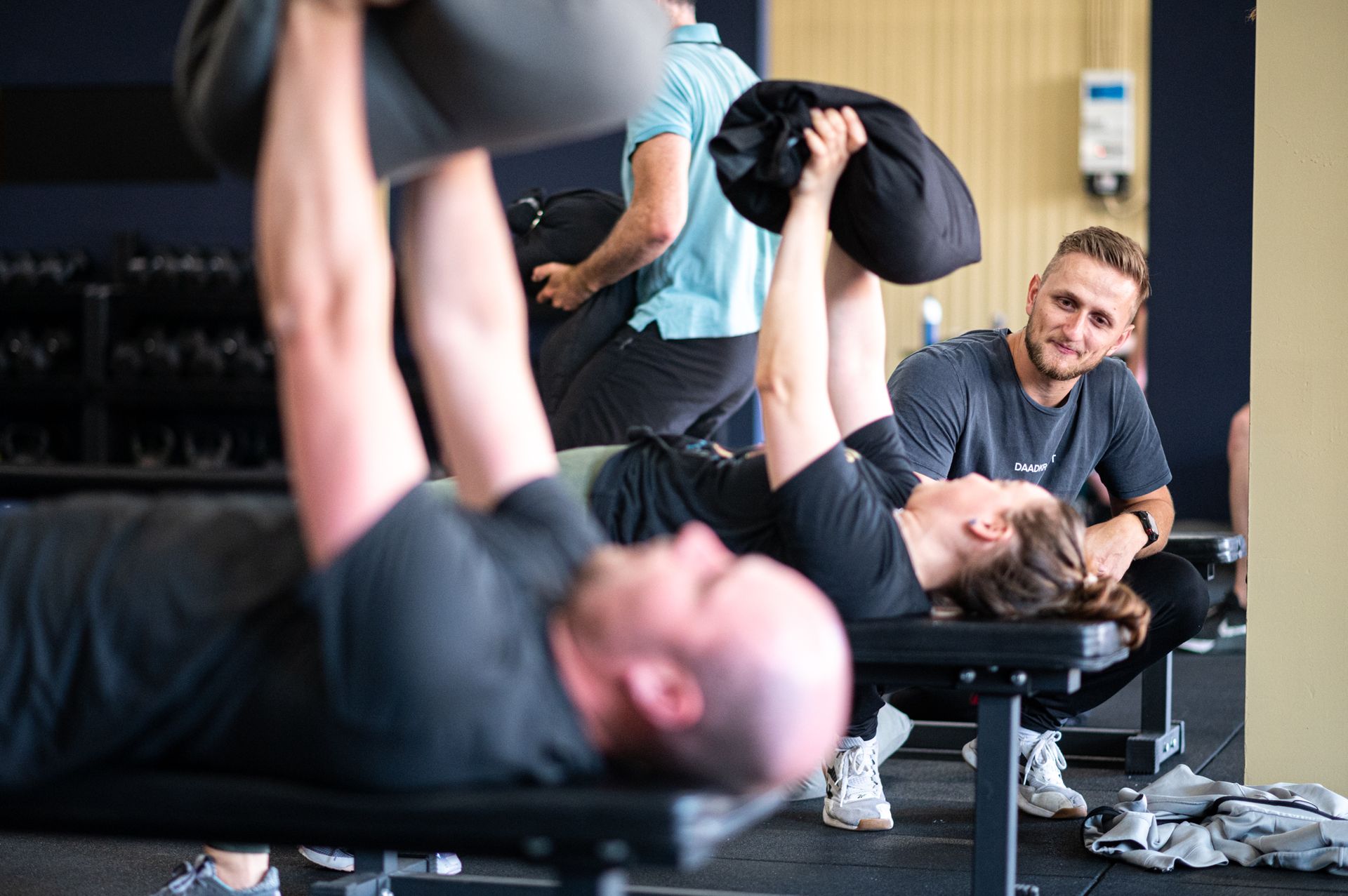 Twee mensen liggen op banken in een sportschool en tillen zandzakken; een trainer kijkt toe.