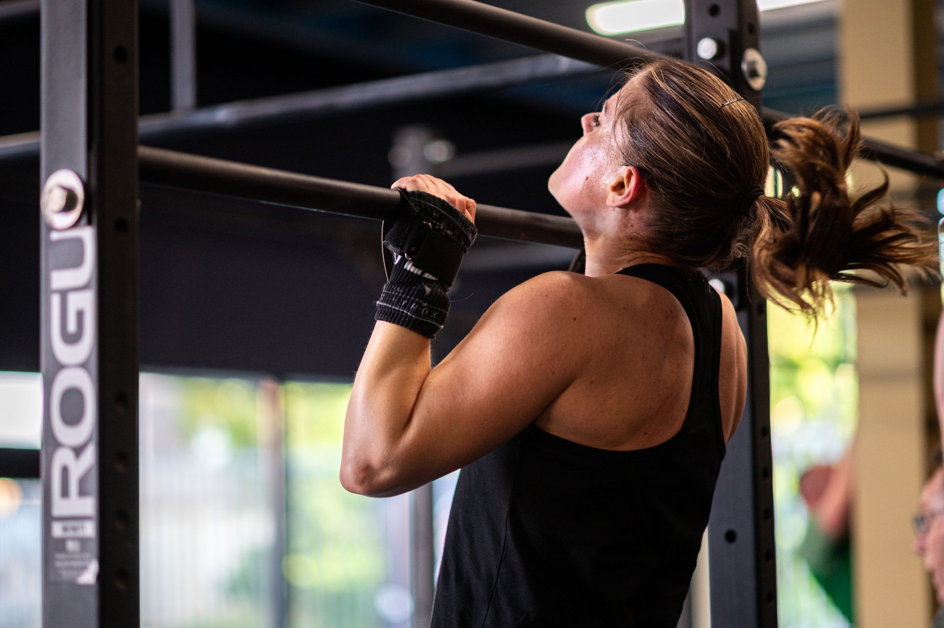 Een vrouw doet een pull-up aan een zwarte stang in een sportschool. Ze draagt een zwart hemdje en handschoenen en lijkt zich flink in te spannen.