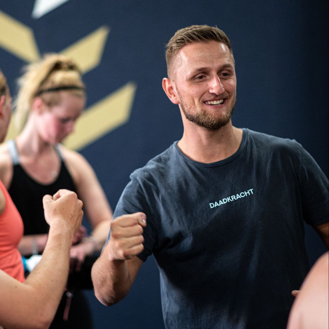 Een man in een donker T-shirt glimlacht en gebaart terwijl hij een fitnessles geeft. Anderen dragen sportkleding.