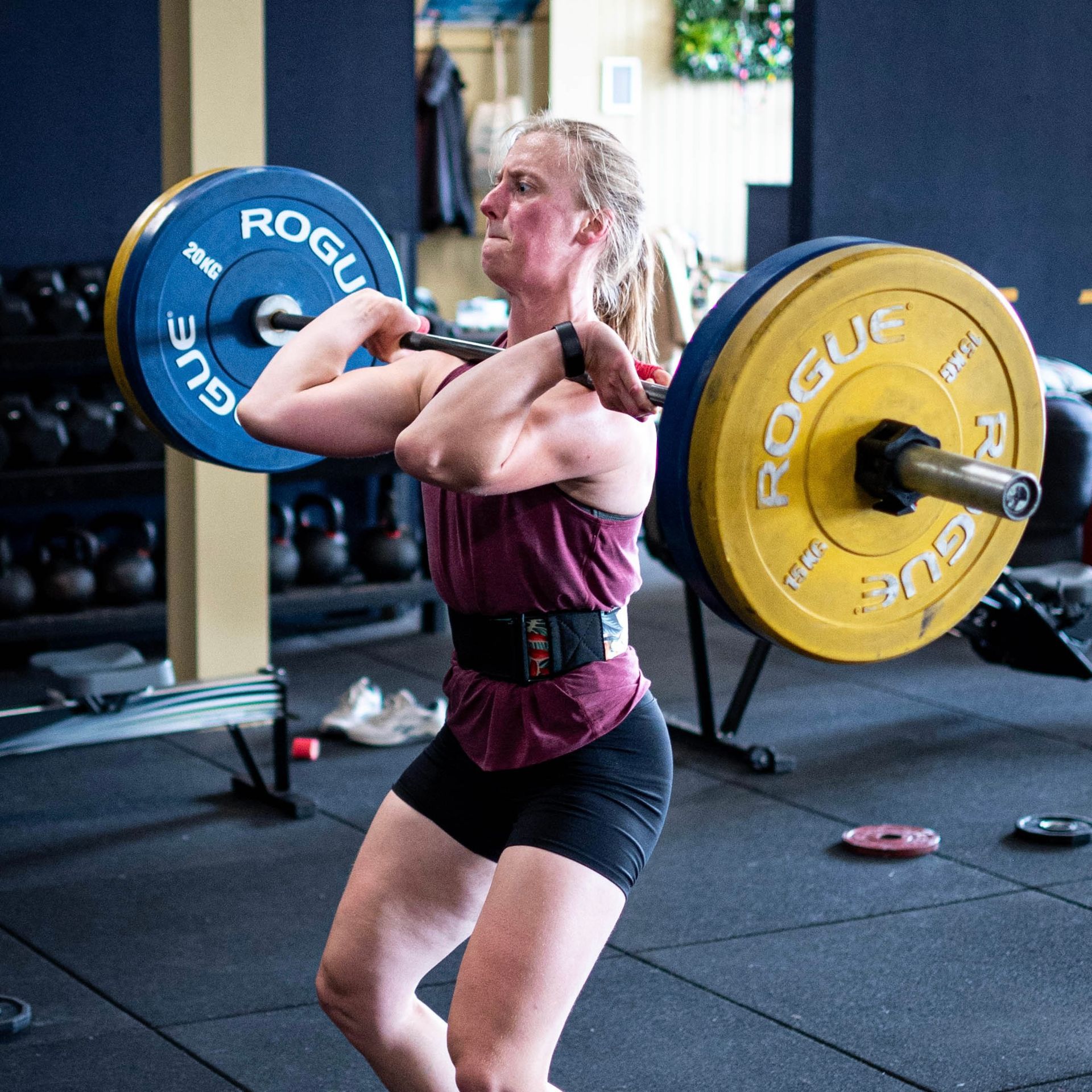 Een vrouw in de sportschool doet een front squat met een halter. Ze draagt een gewichthefgordel en ziet er geconcentreerd uit.