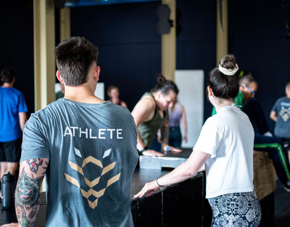 Mensen in een sportschool; twee op de voorgrond kijken weg. Eén draagt ​​een shirt met de tekst 
