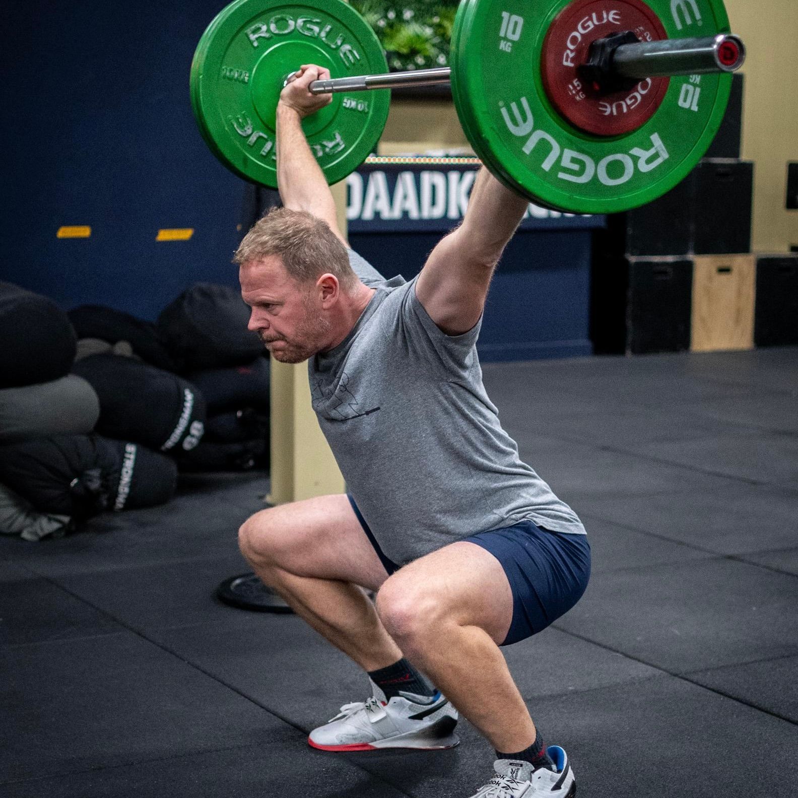 Man in de sportschool doet een squat met een halter boven zijn hoofd. Groene gewichtsschijven, grijs shirt en blauwe korte broek.