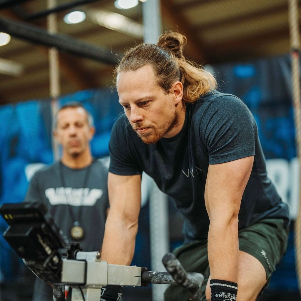 Een man met een knotje traint op een hometrainer; een coach kijkt toe op de achtergrond.