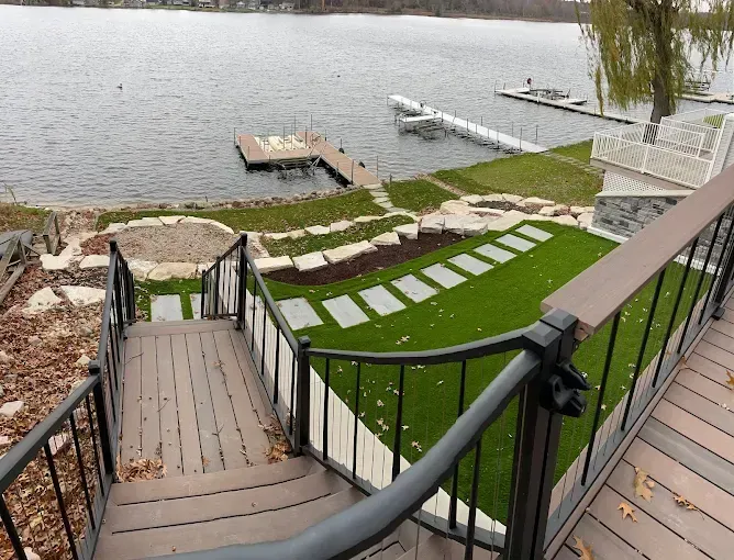 A view from a wooden deck looking down steps to a lakeside yard with artificial turf, stone paths, and two boat docks.