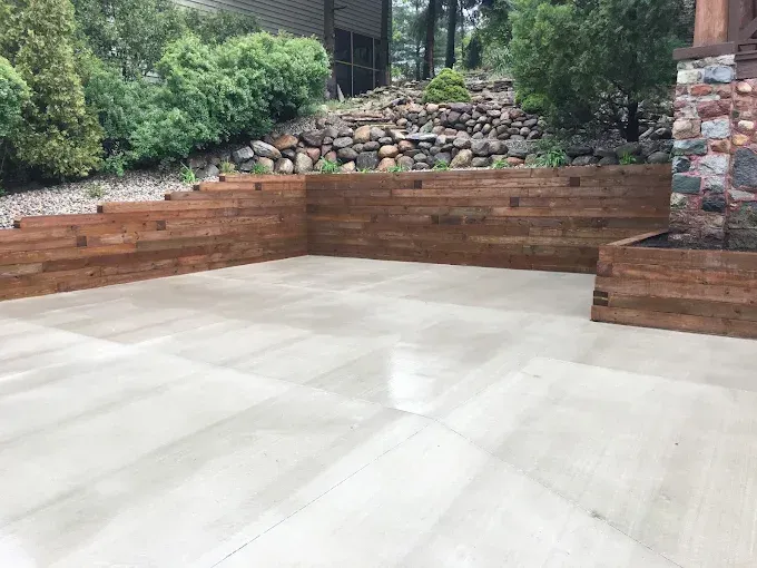 A new concrete patio with tiered wooden retaining walls and a landscaped stone hillside in a residential backyard.