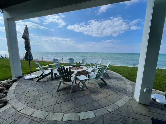 A stone patio with Adirondack chairs around a fire pit, overlooking a calm ocean under a blue, cloudy sky.