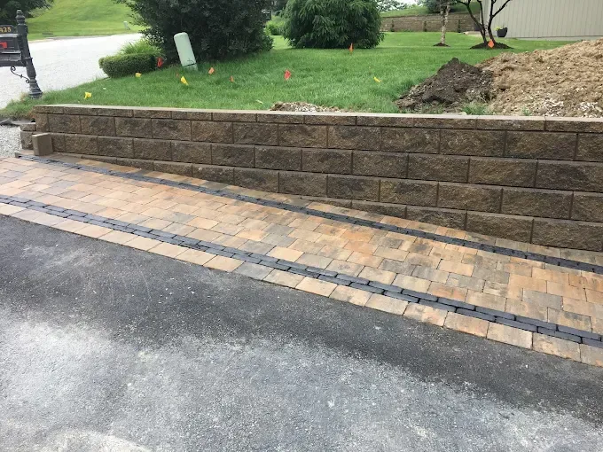 A retaining wall made of tan stone blocks bordering a paved walkway and a driveway with a black drainage grate.
