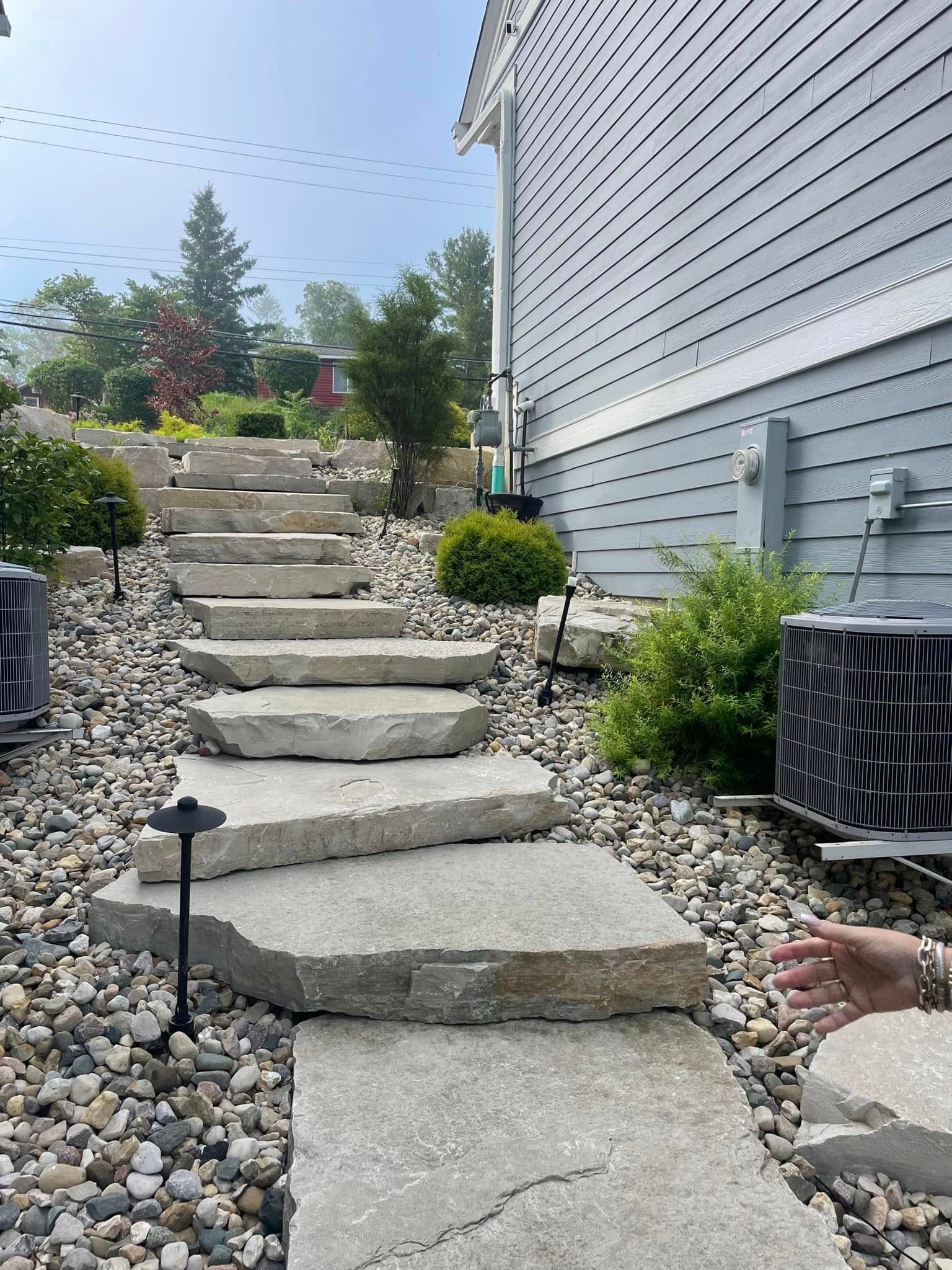 Stone steps set into a sloped landscape next to the gray siding of a house with small shrubs and landscaping lights.