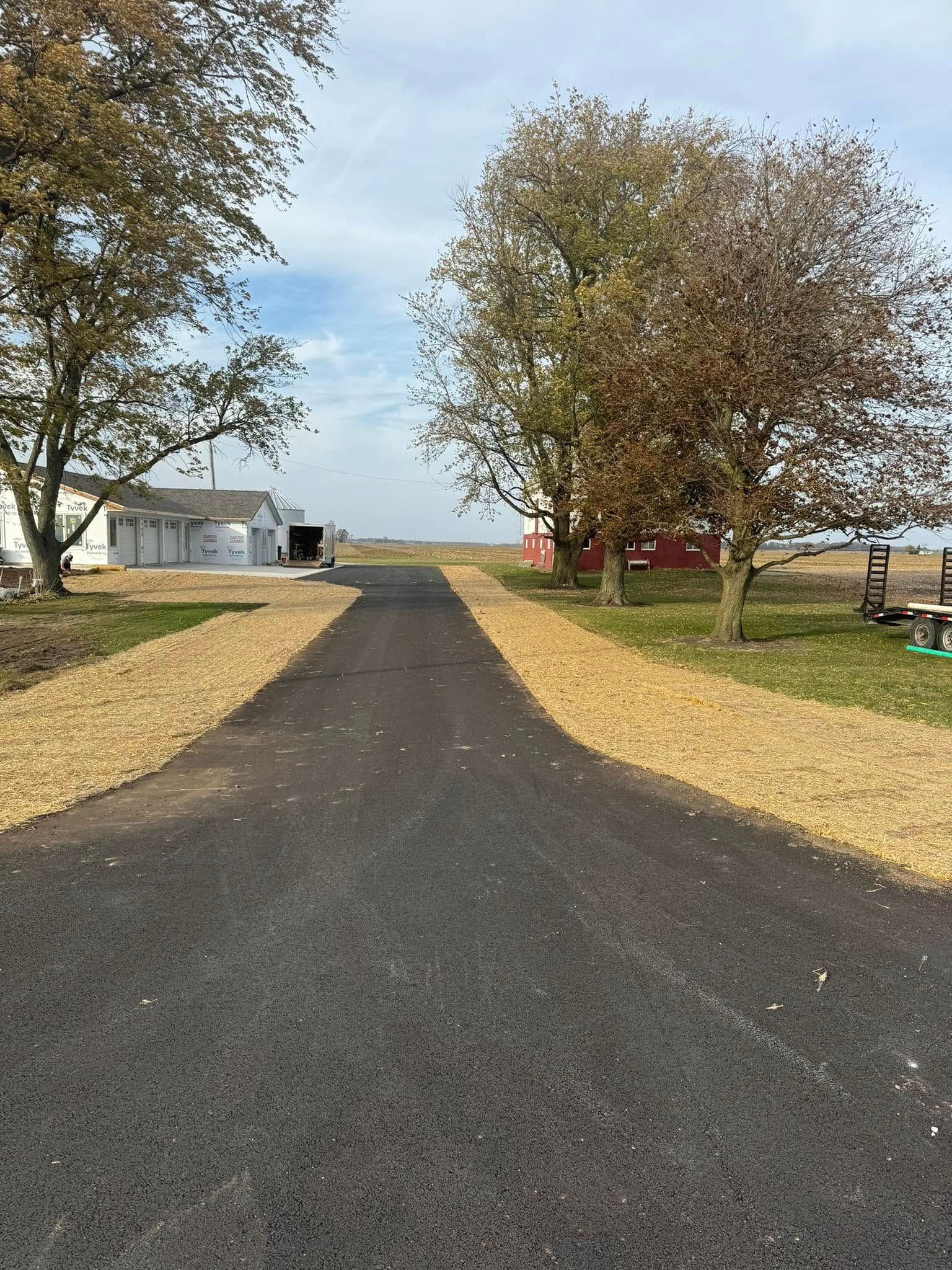 A paved driveway leads toward a white building and a red barn, flanked by trees and gravel borders under a blue sky.