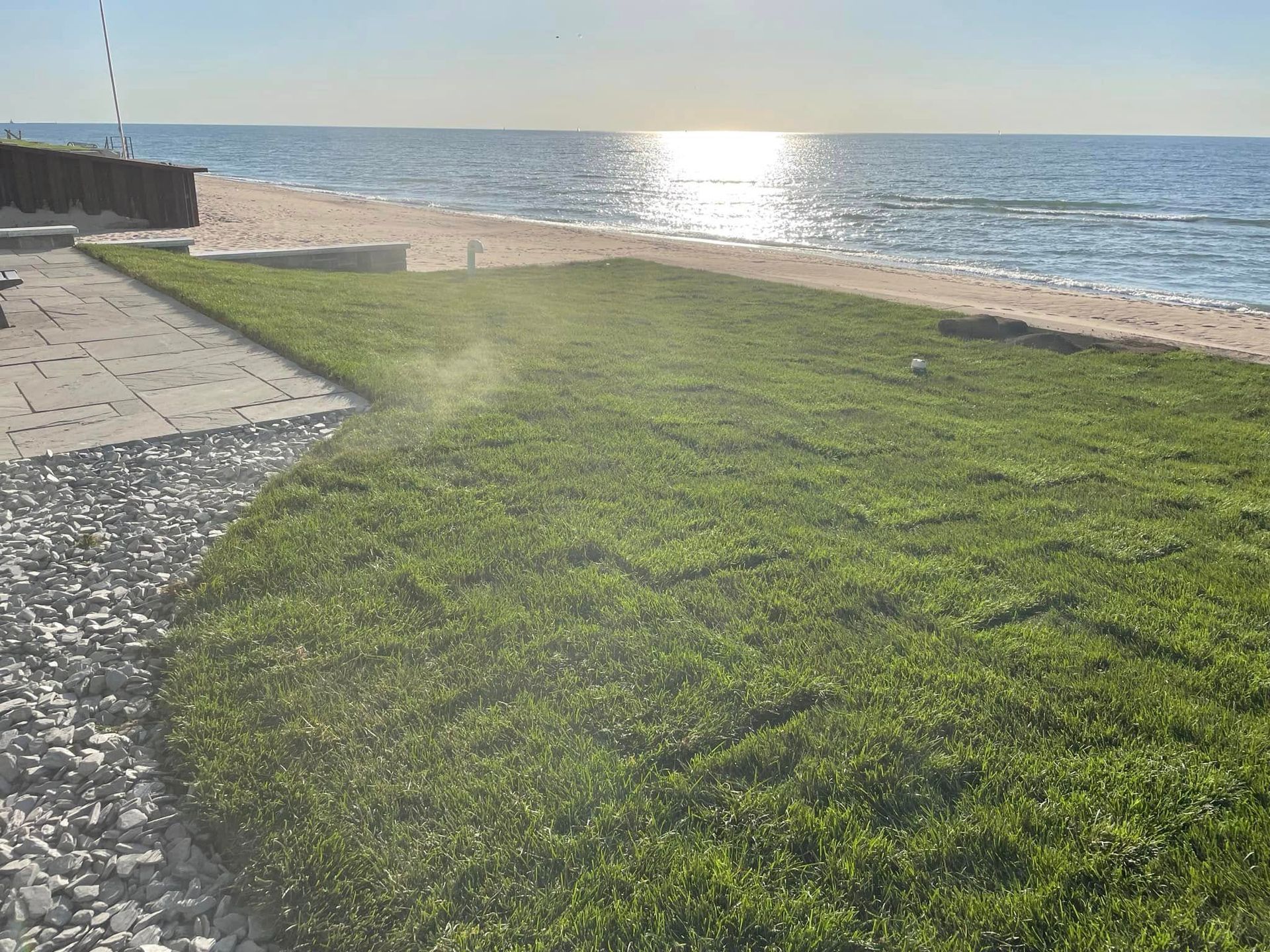 A newly laid patch of green grass lawn bordering a sandy beach and the ocean under a bright, sunny sky.