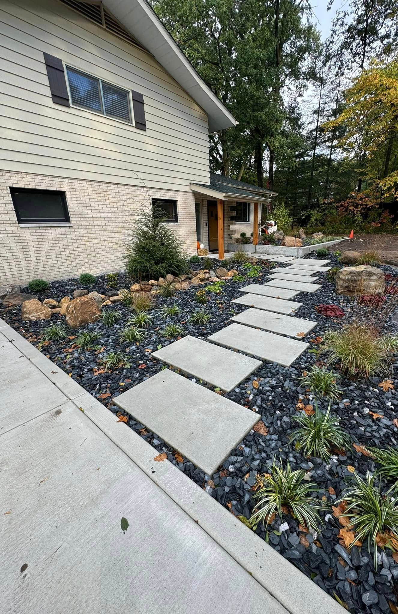 A modern walkway of rectangular concrete pavers set in dark gravel leads to the wooden entrance of a light-sided house.