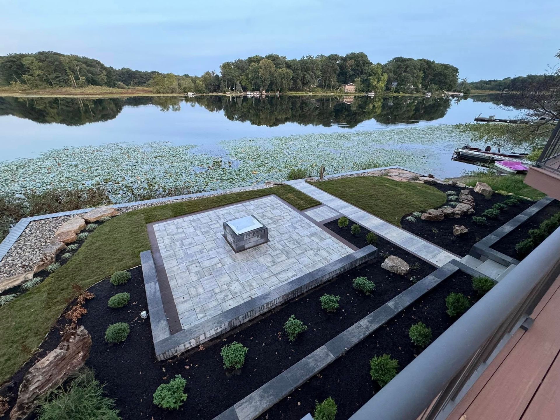 A paved patio with a central square fire pit overlooking a lake with lily pads at dusk, seen from an elevated deck.