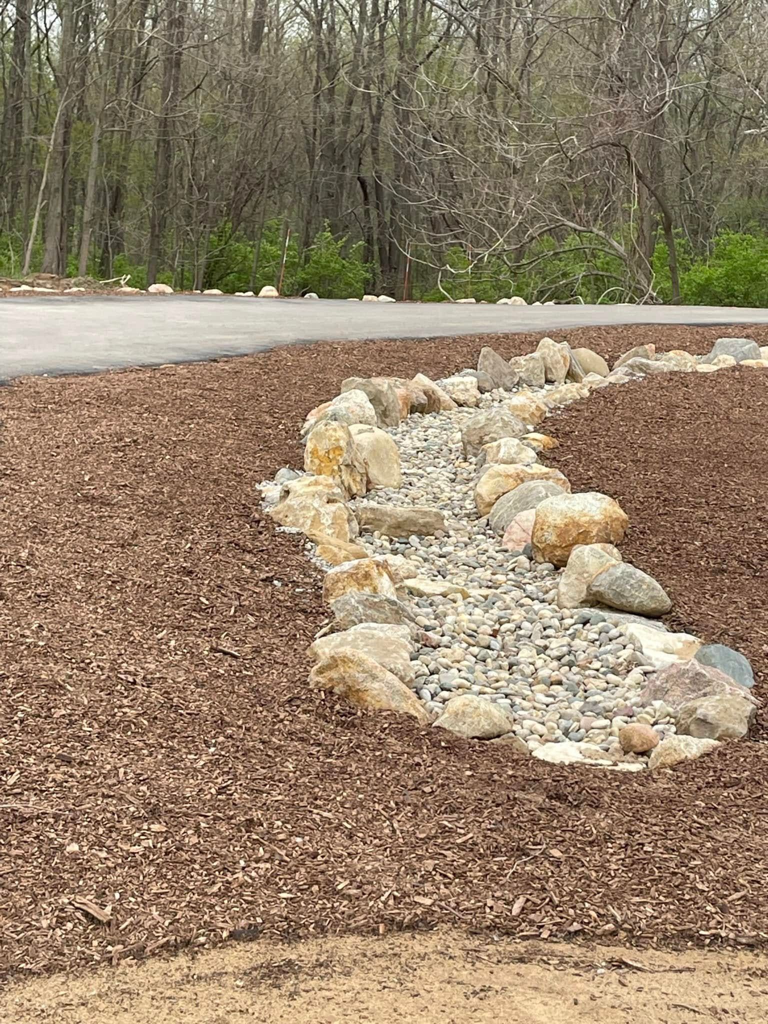 A curved drainage ditch lined with river rocks and bordered by large landscaping boulders in a mulched yard.