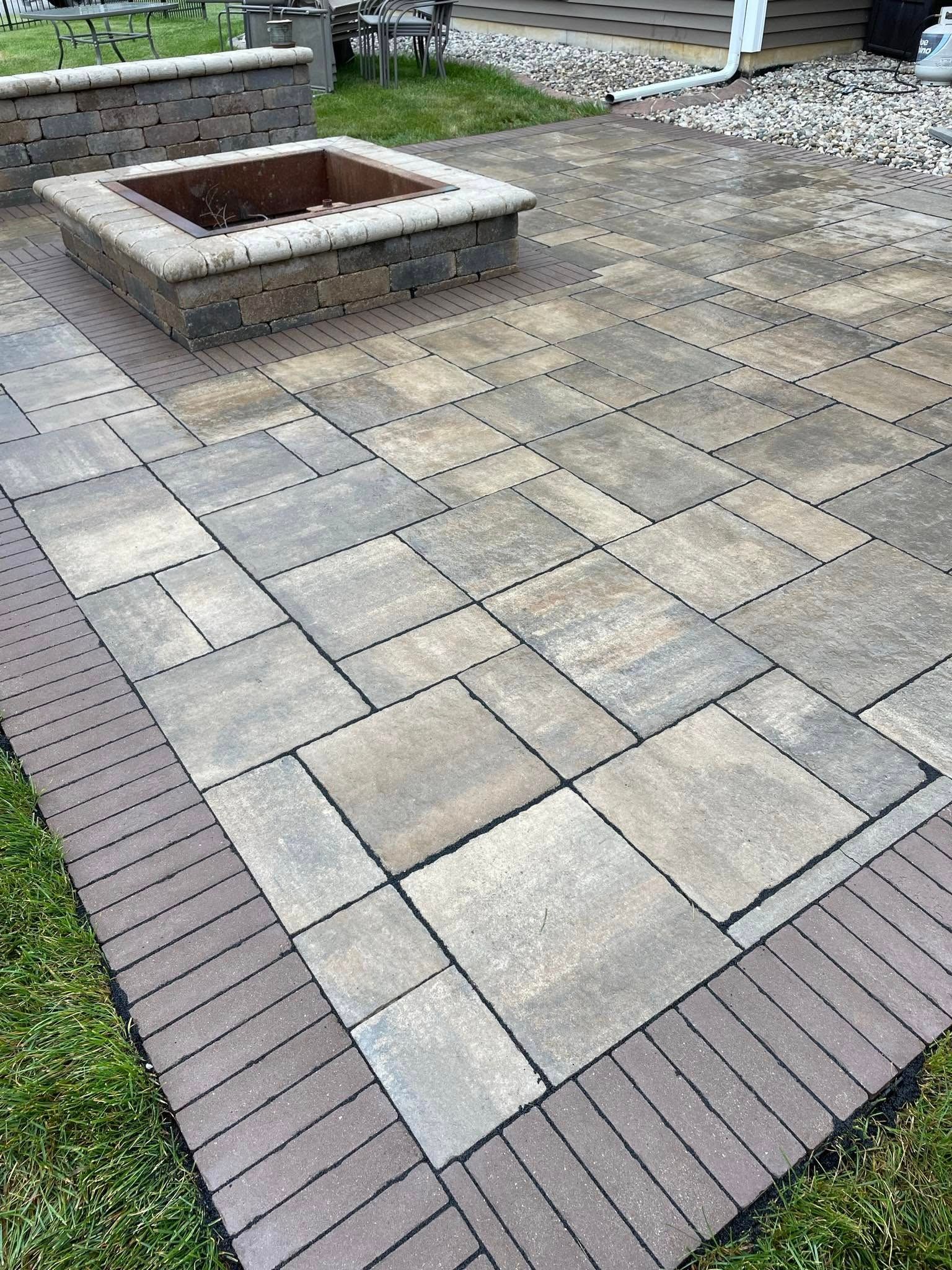 A paved stone patio with a square fire pit, bordered by thin, dark brown bricks on a grassy lawn.
