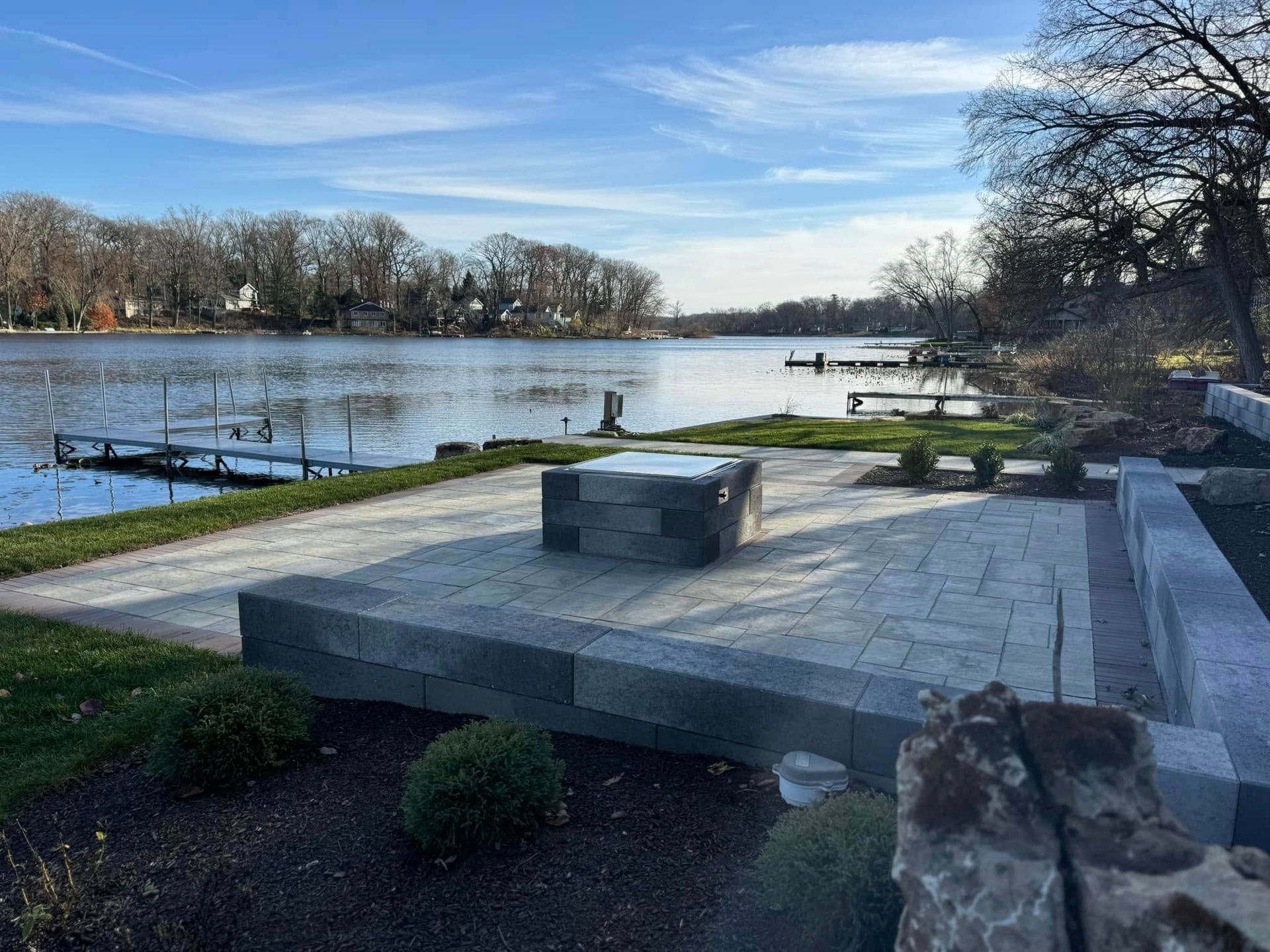 A paved patio with a stone fire pit overlooks a calm lake and distant trees under a blue sky.
