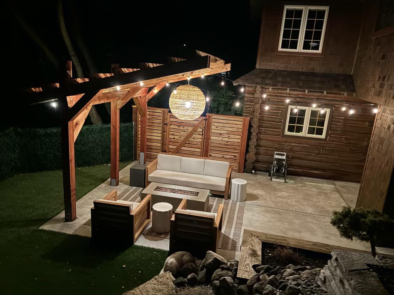 A cozy outdoor patio at night featuring a wooden pergola, string lights, a rattan sphere lamp, and comfortable seating.