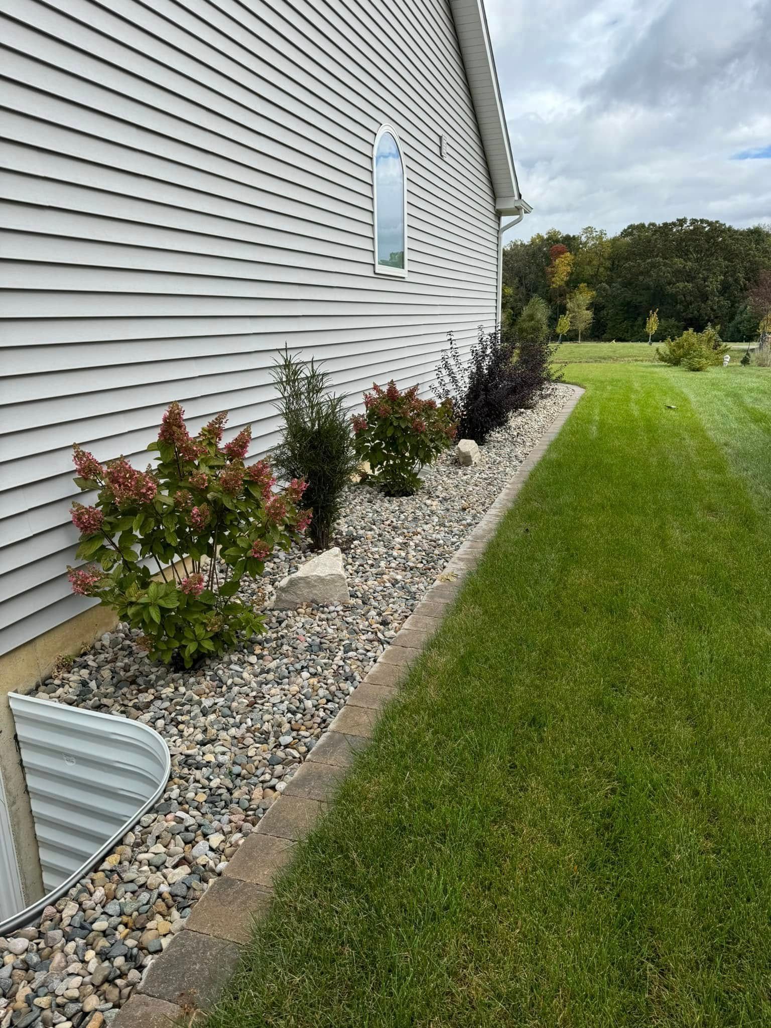 Light grey house siding with a stone-filled garden bed, several green shrubs, and a metal window well along a grassy lawn.