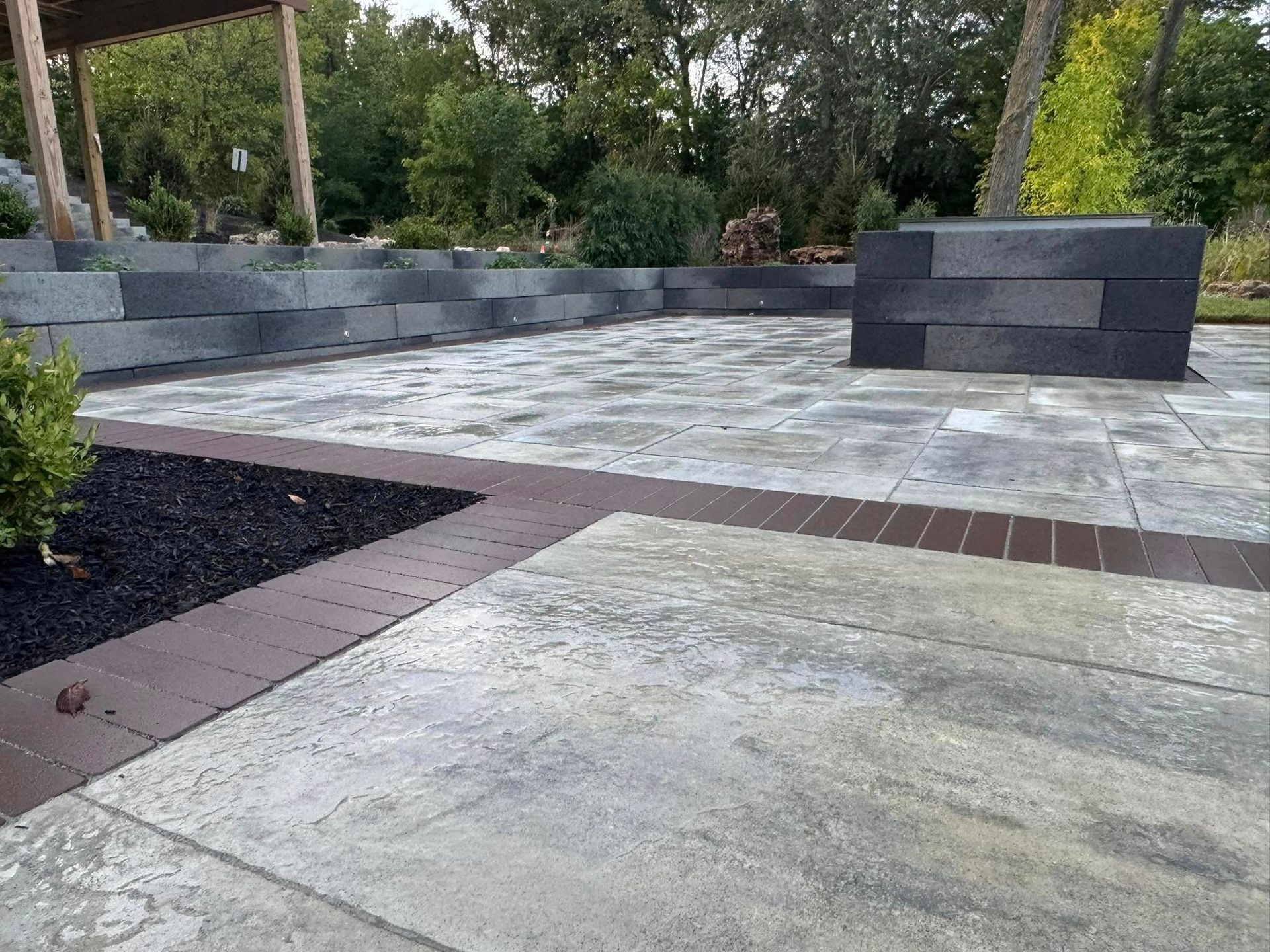A stone patio features light-gray textured pavers bordered by dark-brown bricks, with matching gray block retaining walls.