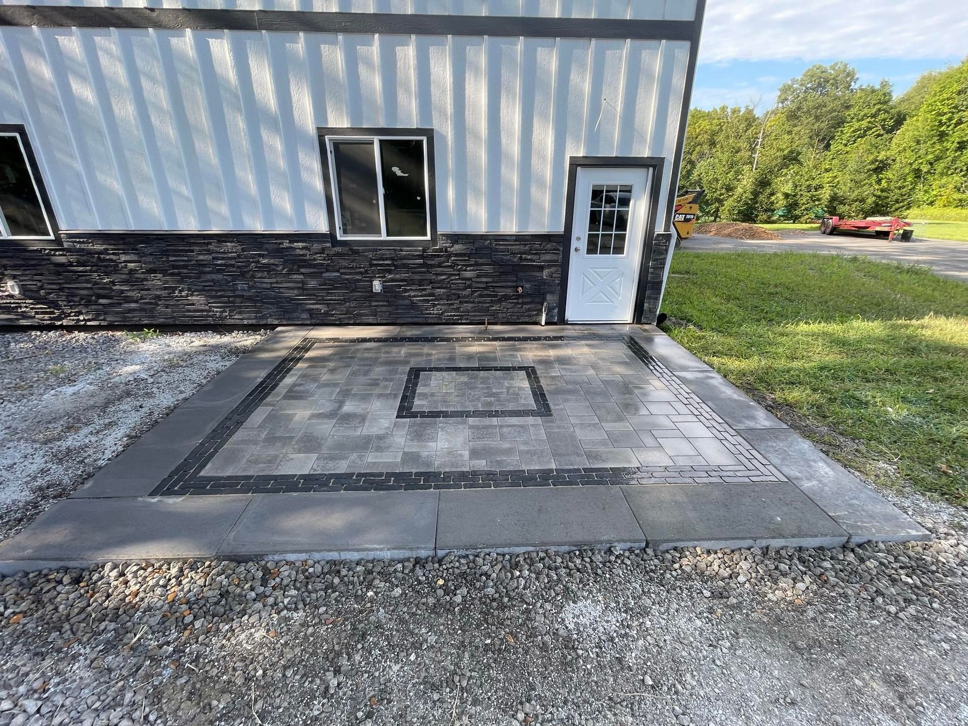 A newly installed gray paver patio with a dark charcoal border and central square inlay outside a white metal building.