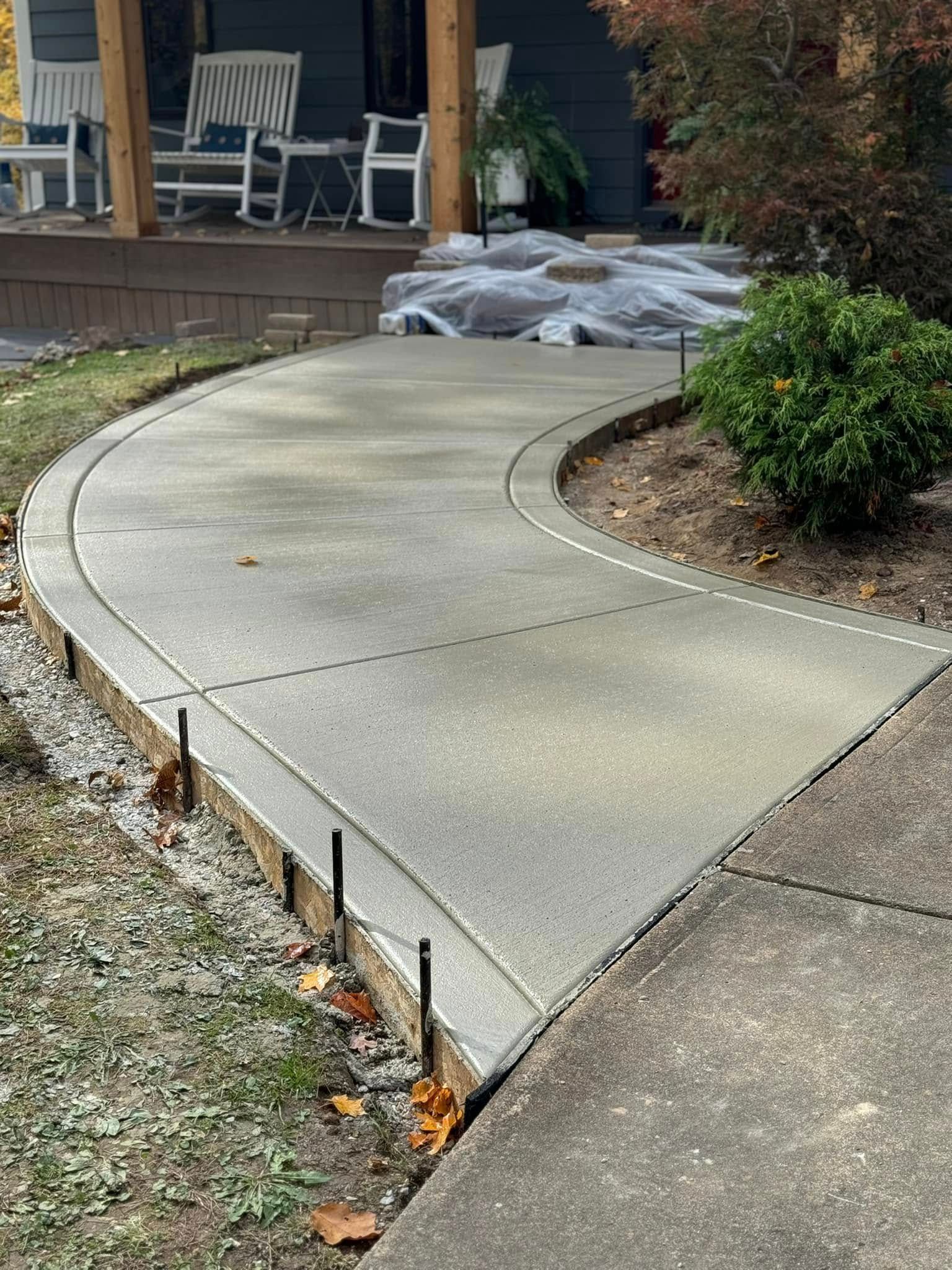 A newly poured concrete walkway curves toward a porch with rocking chairs, outlined by wooden forms and metal stakes.