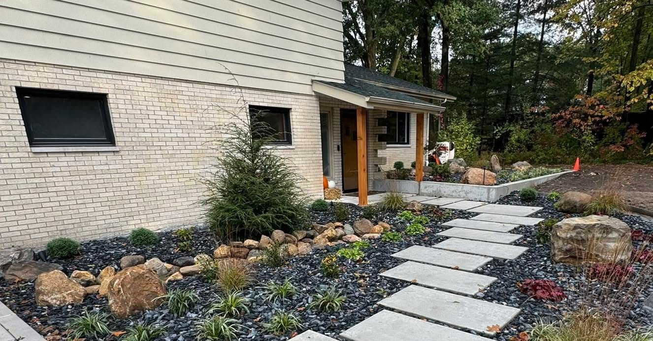 A modern house with cream brick, a stone walkway, and a landscaped front garden with dark mulch and small shrubs.