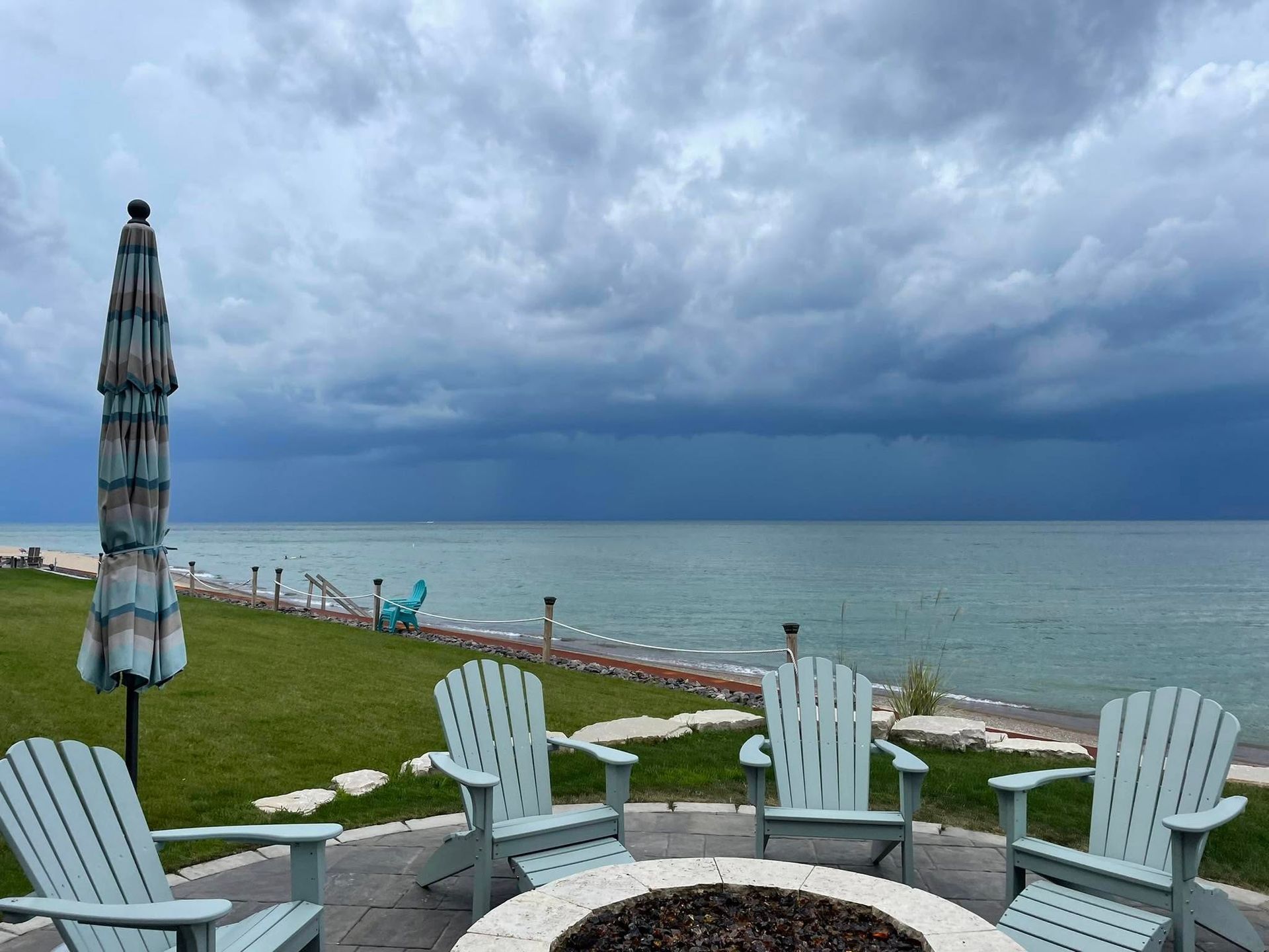 Four light blue adirondack chairs arranged around a circular fire pit on a patio overlooking a stormy lake.