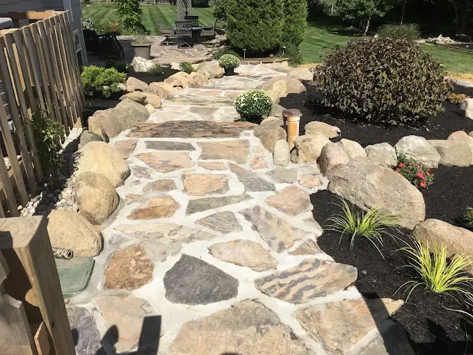 A stone flagstone walkway with mortar joints leads through a landscaped yard bordered by large rocks and greenery.