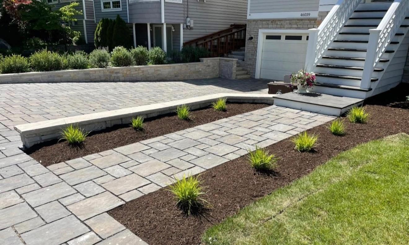Modern driveway with gray pavers, mulch beds, small shrubs, and a white staircase beside a garage